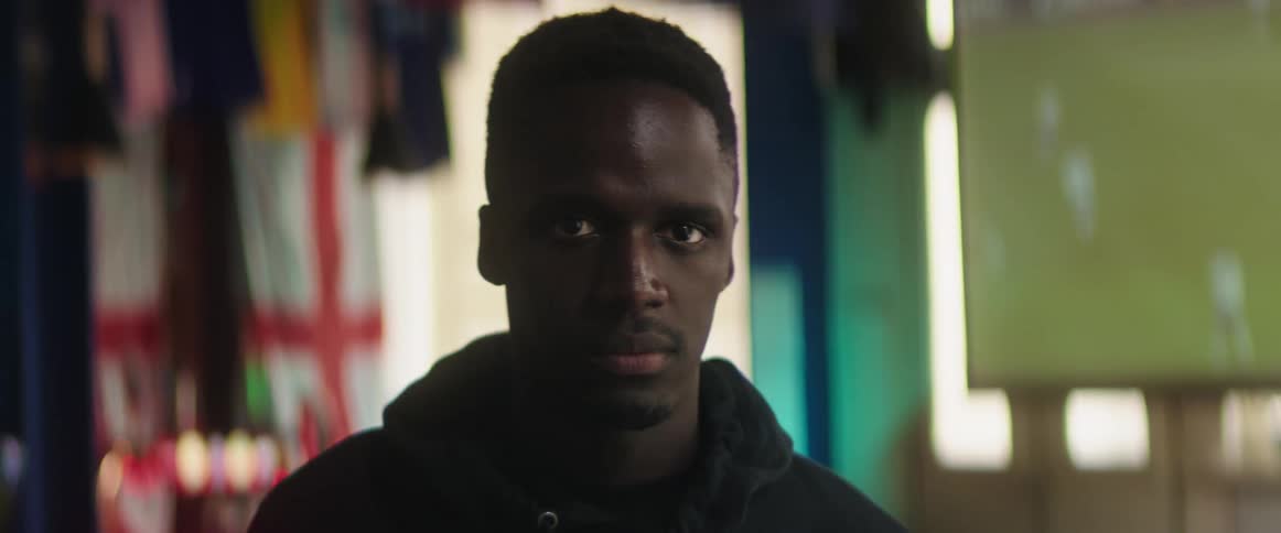 Man with black coat looks at camera smiling in front of television with football match on in bar