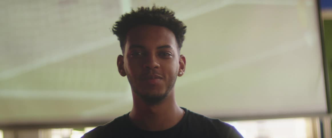 Boy with earring and stubble beard stands in front of television screen looking at camera and smiling