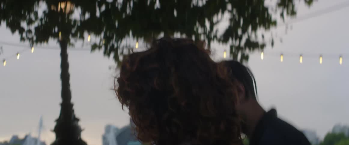 A man and woman stand close and chat as they walk in a park across from downtown