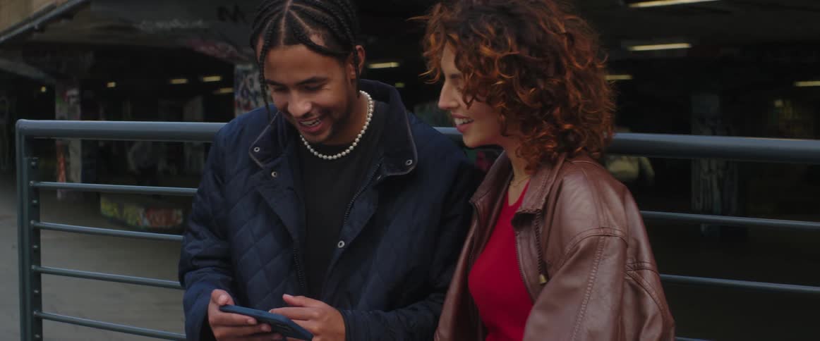 A man and woman stand and talk and laugh and look at a cell phone as teenager skateboard behind them and people walk