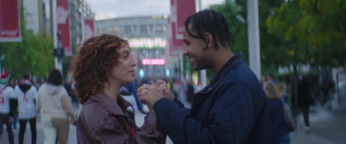 A man and woman stand on a busy street and hold hands and talk as many people walk past