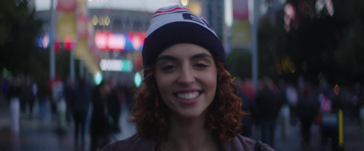 Woman with beanie and brown wavy hair faces the camera with eye contact laughing in front of football stadium