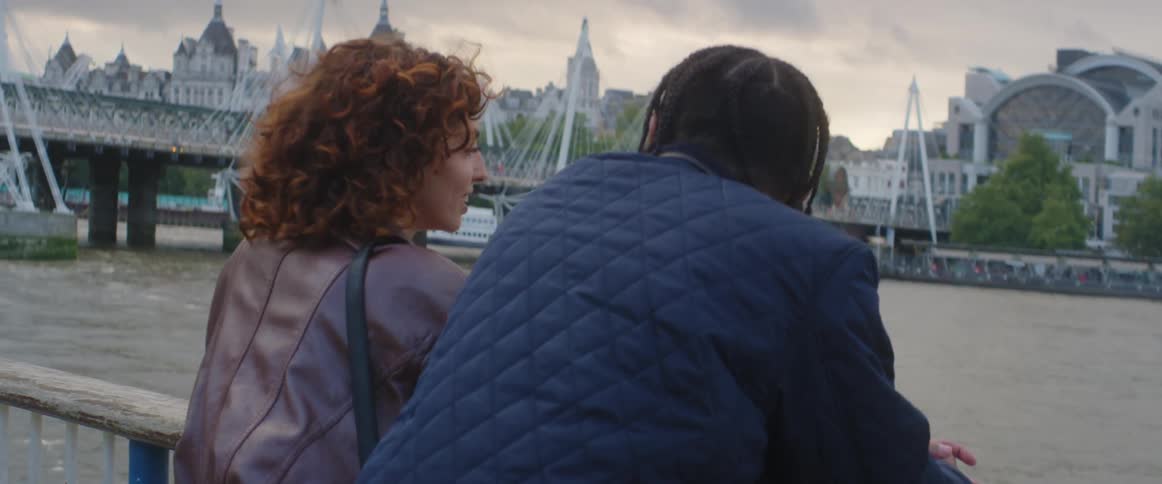 A young couple stand on a bridge in a city having a conversation