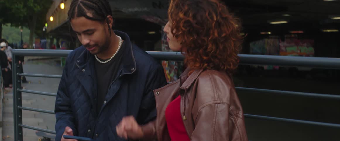 A woman and man are standing just outside a building leaning against a fence talking