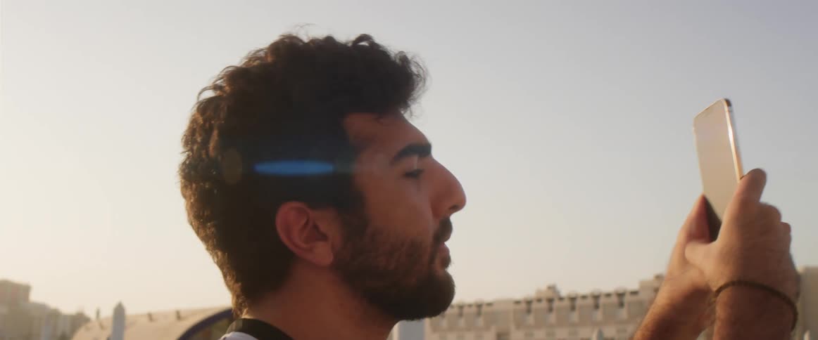 A man is standing near buildings and holding up his cell phone taking a picture