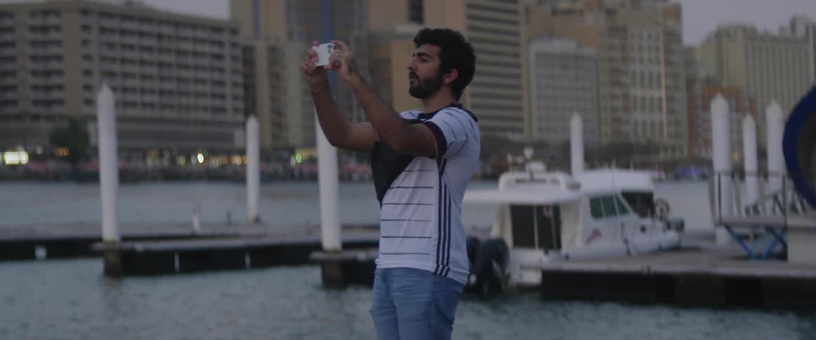 A man stands at a marina and holds up his phone to take a photograph