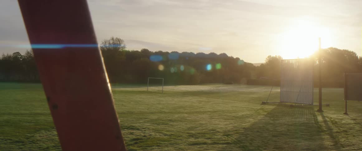 The sun shines brightly upon a soccer goal on a field surrounded by trees