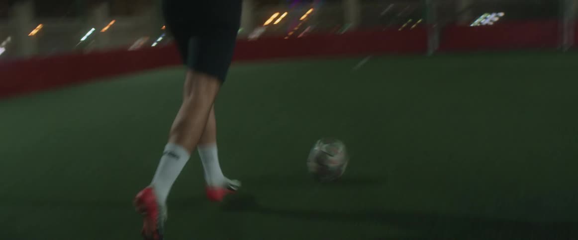 Man dribbles soccer ball across sports field to goal at night with city buildings in background