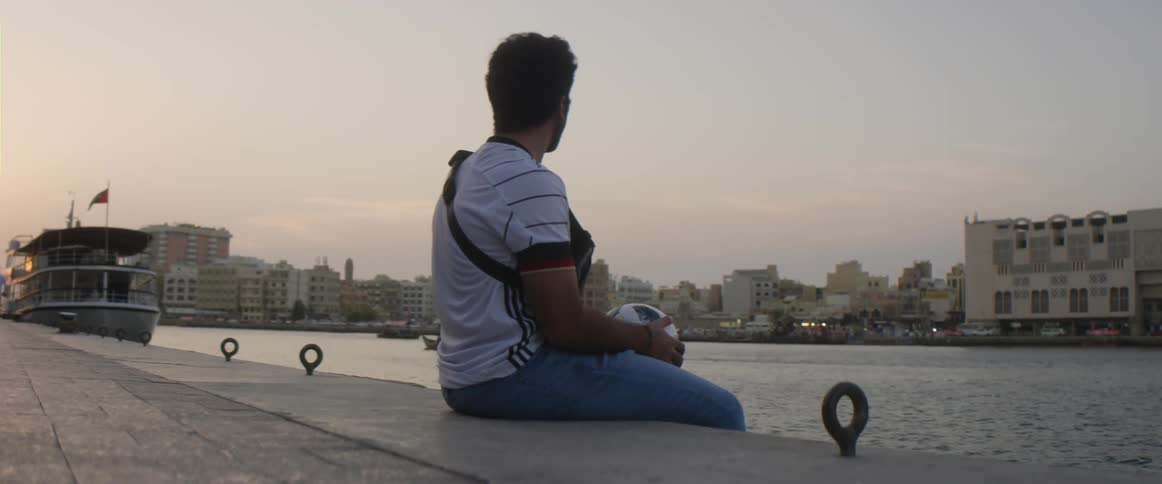A man is sitting by a river and holding a soccer ball