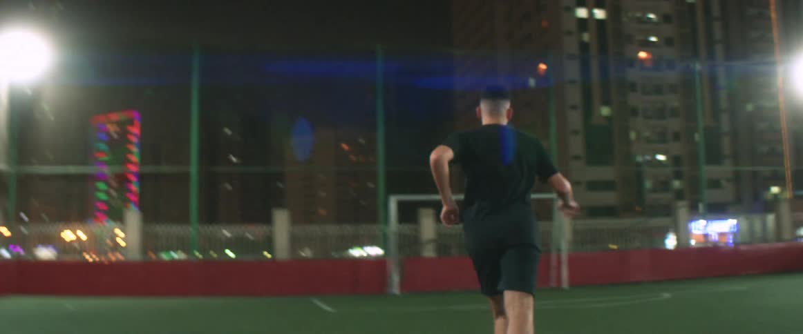 Youth practices soccer alone on field at night with buildings in background