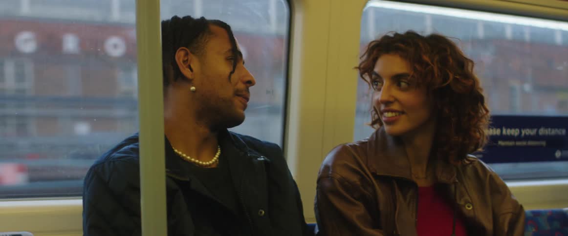 Man with braids earrings necklace and black leather jacket talks with woman with brown wavy hair and leather jacket on traveling train