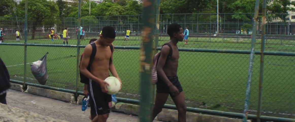 Boys with ball walk along sidewalk past soccer field where team is playing