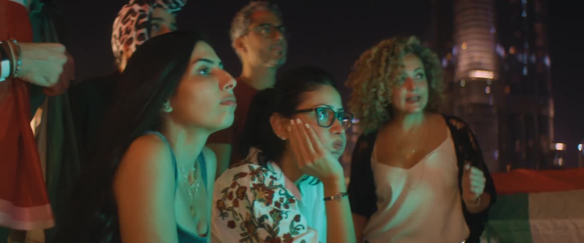 A group of friends sit and stand on a roof top watching something and shaking their heads and clenching their fists