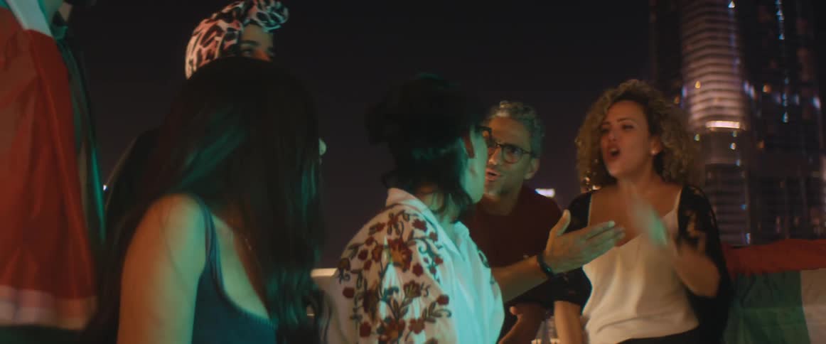 A group of men and women stand and sit on a rooftop and look off in to the distance as one woman raises her arms and points and argues with a man and another woman claps
