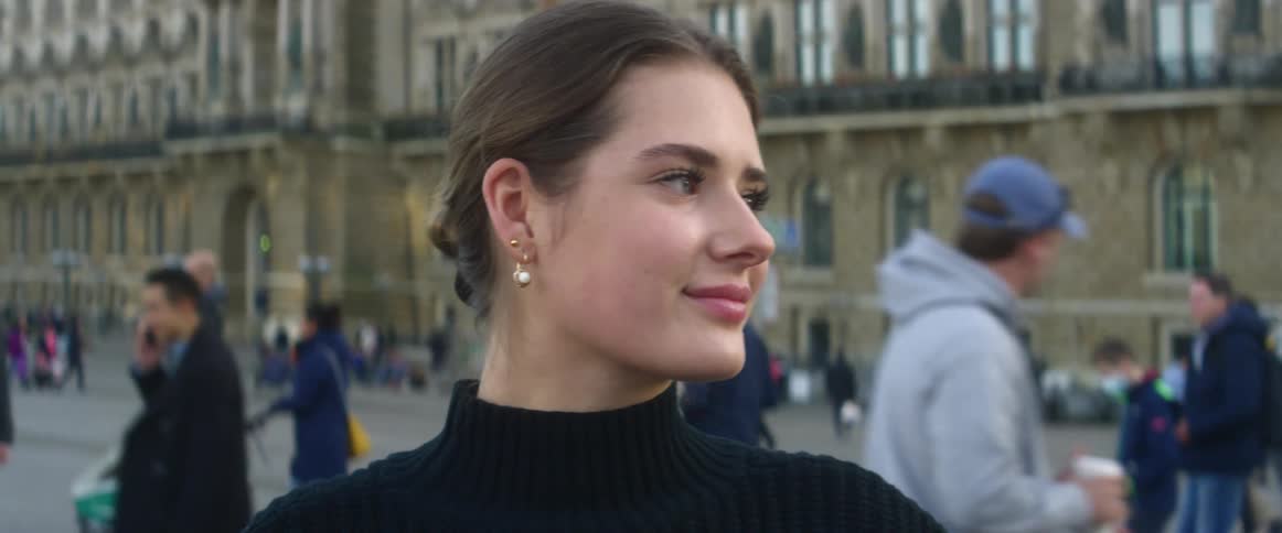 Woman with brown hair earrings and black sweater faces the camera with eye contact in city square filled with people
