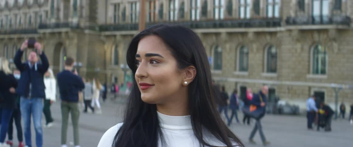 A smiling woman stands facing the camera with a building and people in the background