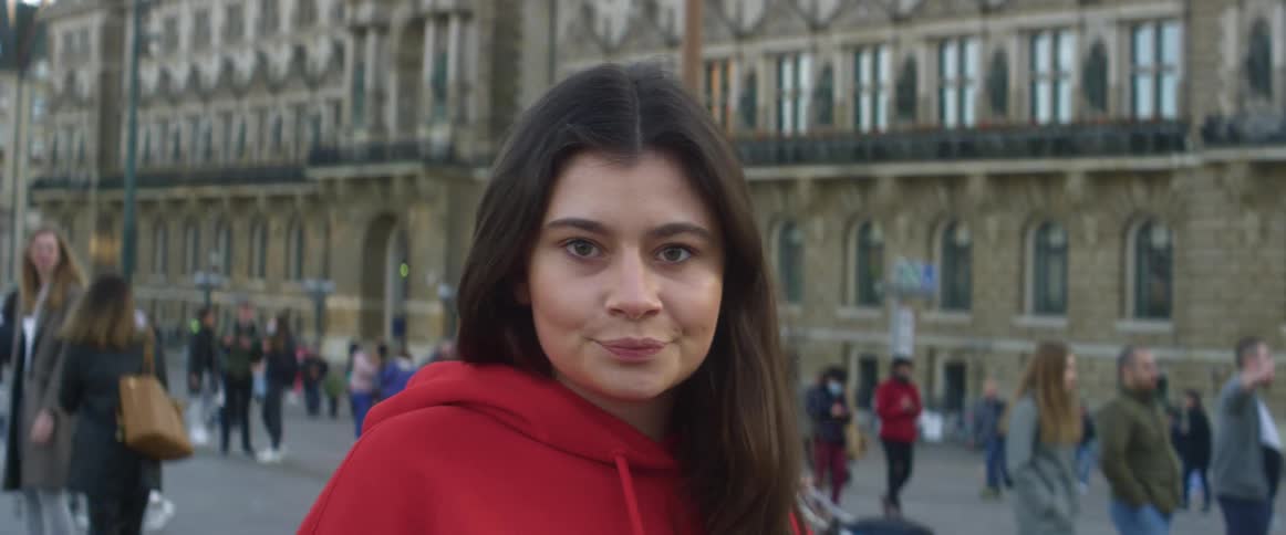 Woman with red hoodie and brown hair faces the camera with eye contact in city square filled with people