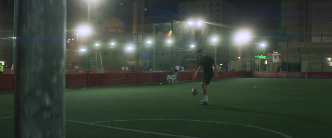 A lone man practices dribbling and shooting a soccer ball at night in the middle of a city in an enclosed field