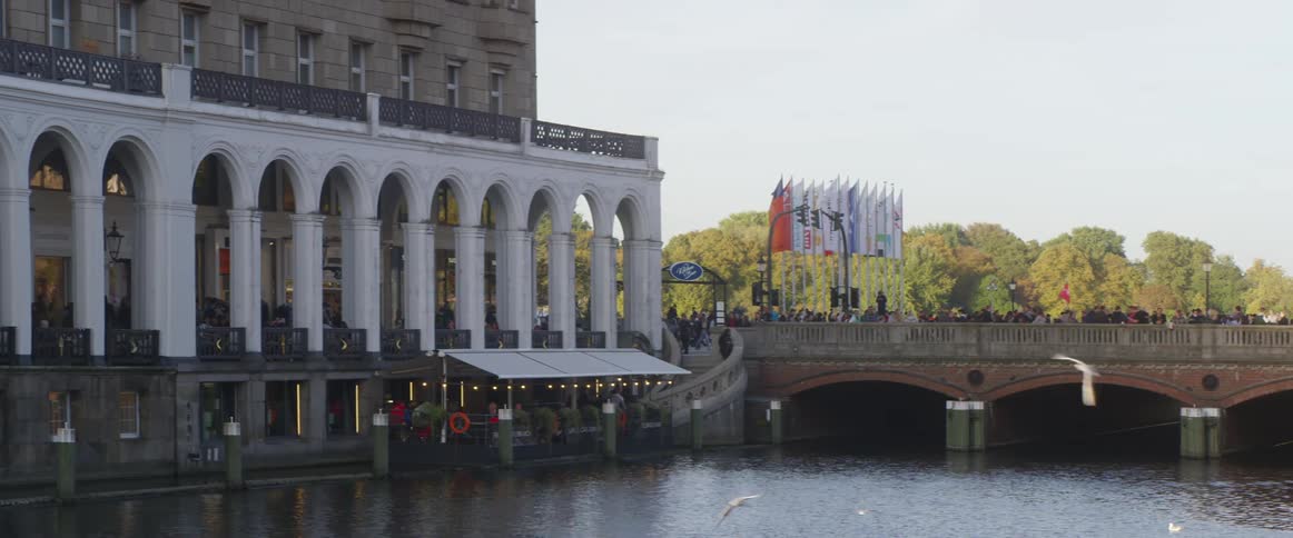 Birds fly over river as people mill along bridge and nearby building decorated with a series of arches
