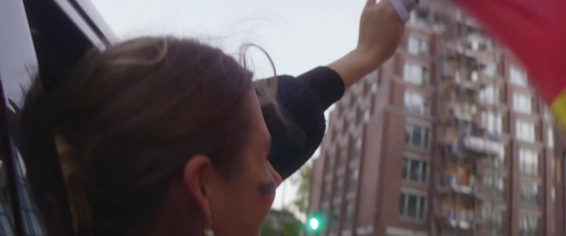 A woman holding a flag hangs out a car window as it drives down a city street and cheers