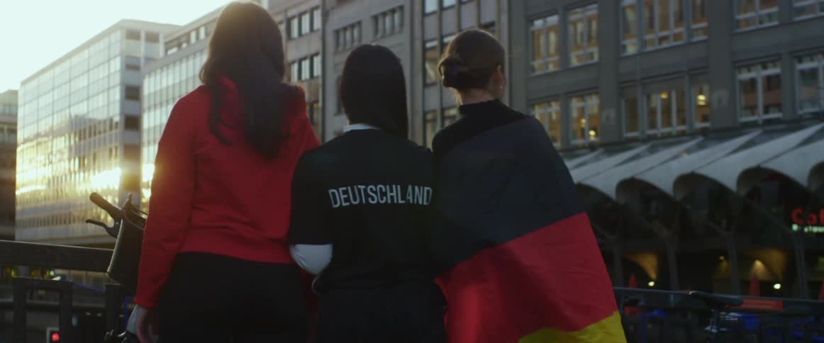 Three teenage girls stand arm in arm on a bridge in a city and talk and laugh