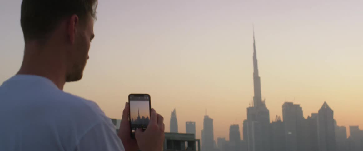 A man holds his cell phone taking photos of the low sun behind tall city buildings