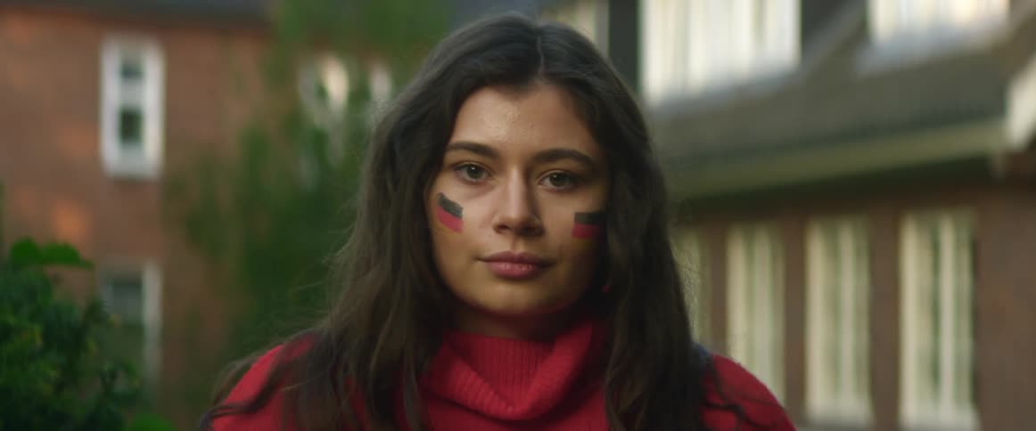 Womanwith face paint on cheeks stands outdoors with building in background fixes her hair and smiles slightly