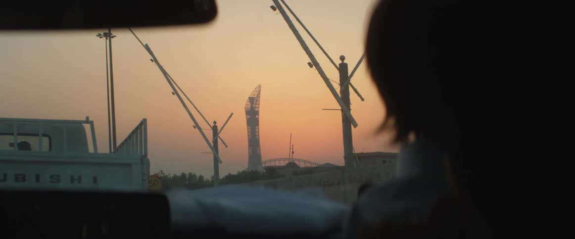 Person sits in vehicle near long armed lights with bridge and red sky in background