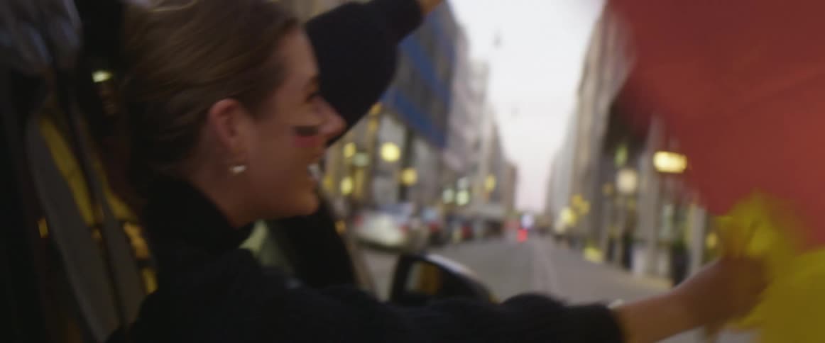 A woman holds a german flag out the passenger side window of a vehicle as it travels down a street in a metropolis during the day