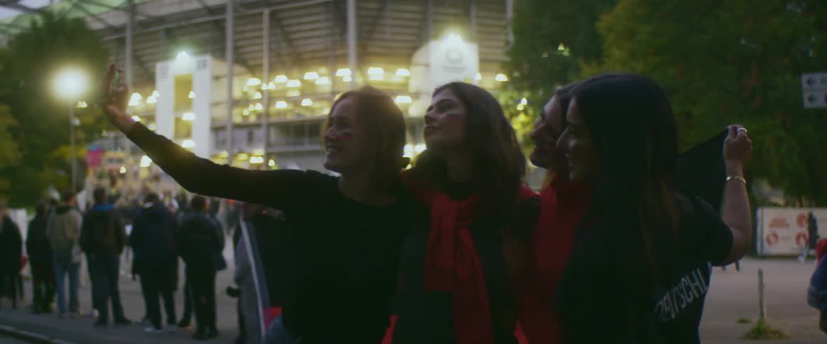 Friends take selfie with a smartphone while standing in front of a stadium at evening