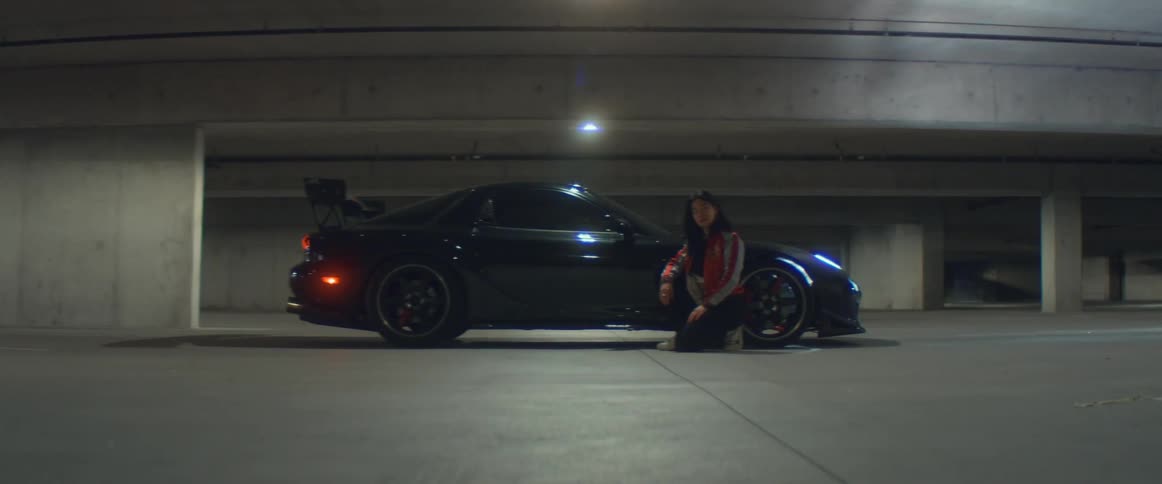 A woman in a race jacket poses next to a sports car in a large parking garage