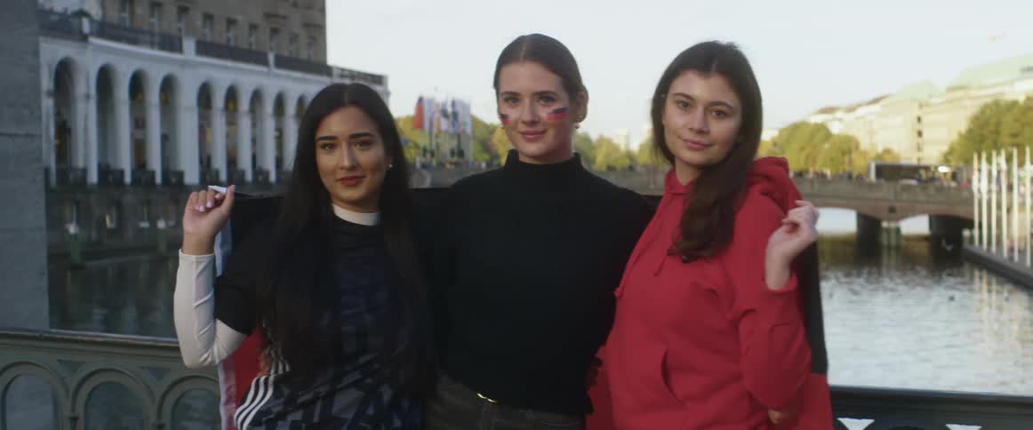 Three women are standing on a bridge over a river posing with a flag draped around them