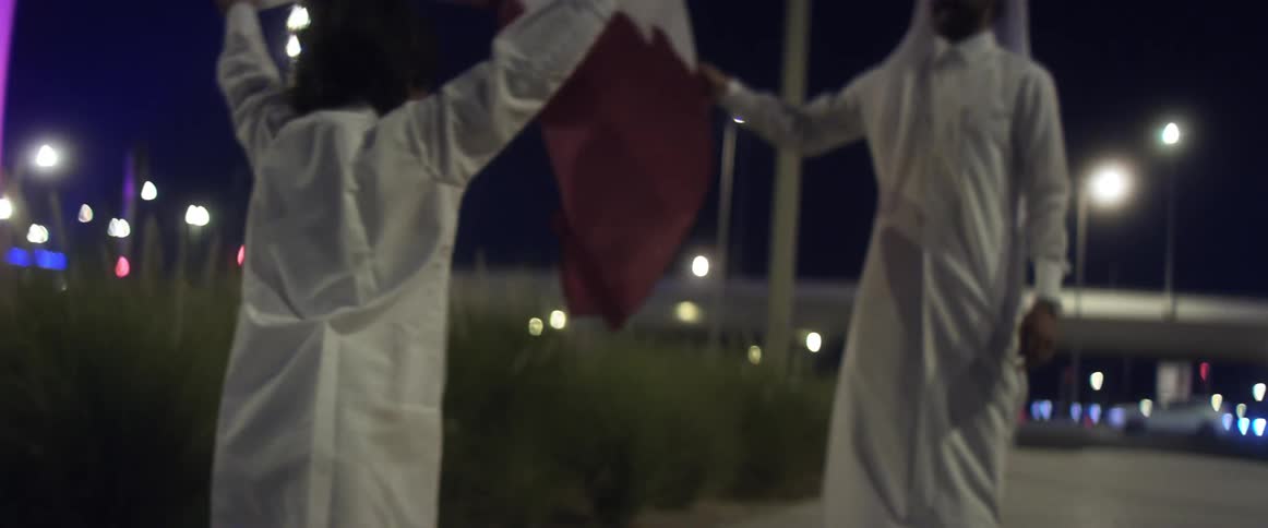 Father and son wave Qatar flag while standing on sidewalk near highway at night