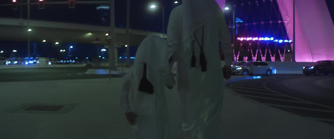 An adult and child in traditional clothing hold hands while walking through a city next to a busy road at night