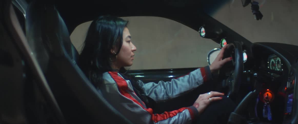 A woman in a racing jacket sits in the drivers seat of a race car parked in a garage