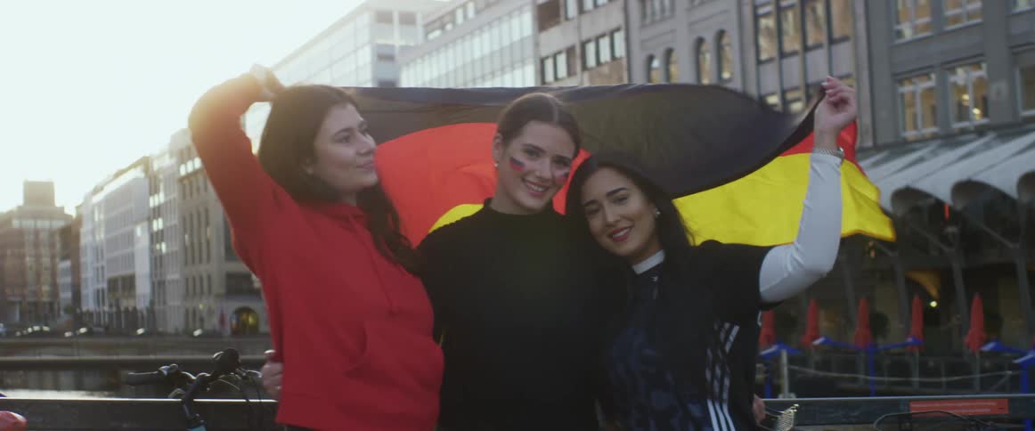 Three women hug and hold a flag over their heads as they pose on a bridge downtown