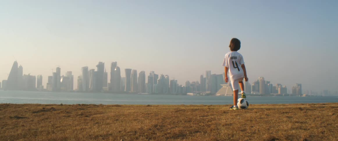 A boy stands with his leg on a soccer ball as he stares across the sea to downtown