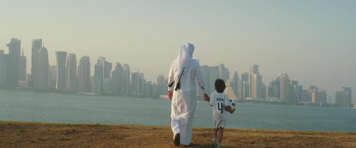 A father in traditional white abaya walks toward the water holding his sons hand