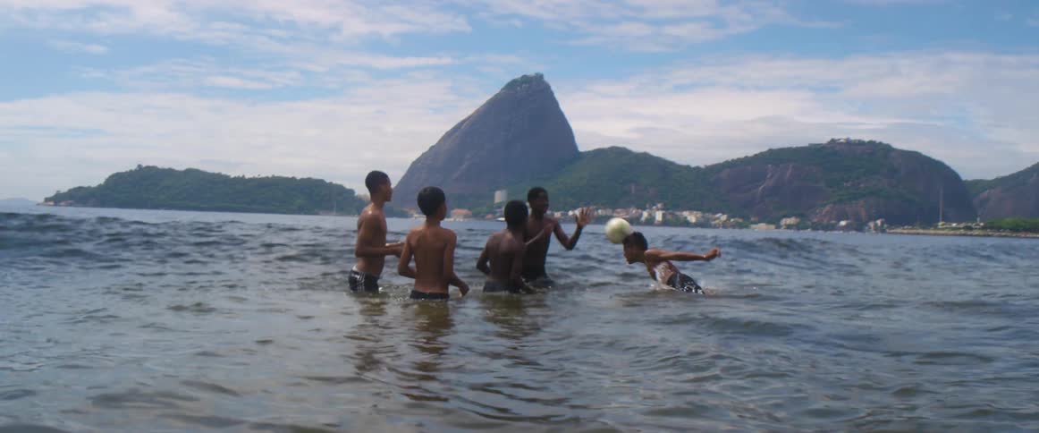 A group of boys standing in the ocean water playing with a soccer ball jump as a wave rolls by