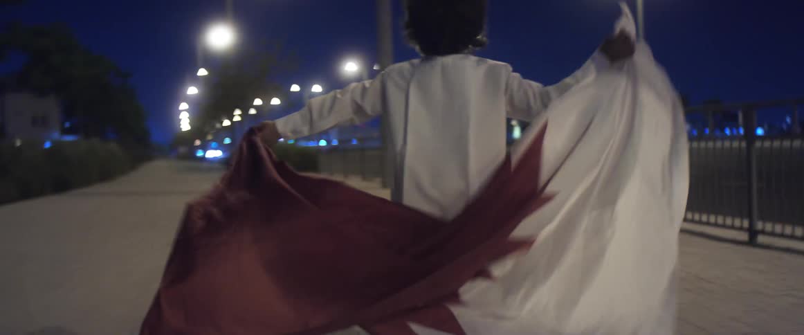 A young boy holds a flag behind his back as he runs along a sidewalk as vehicles drive by