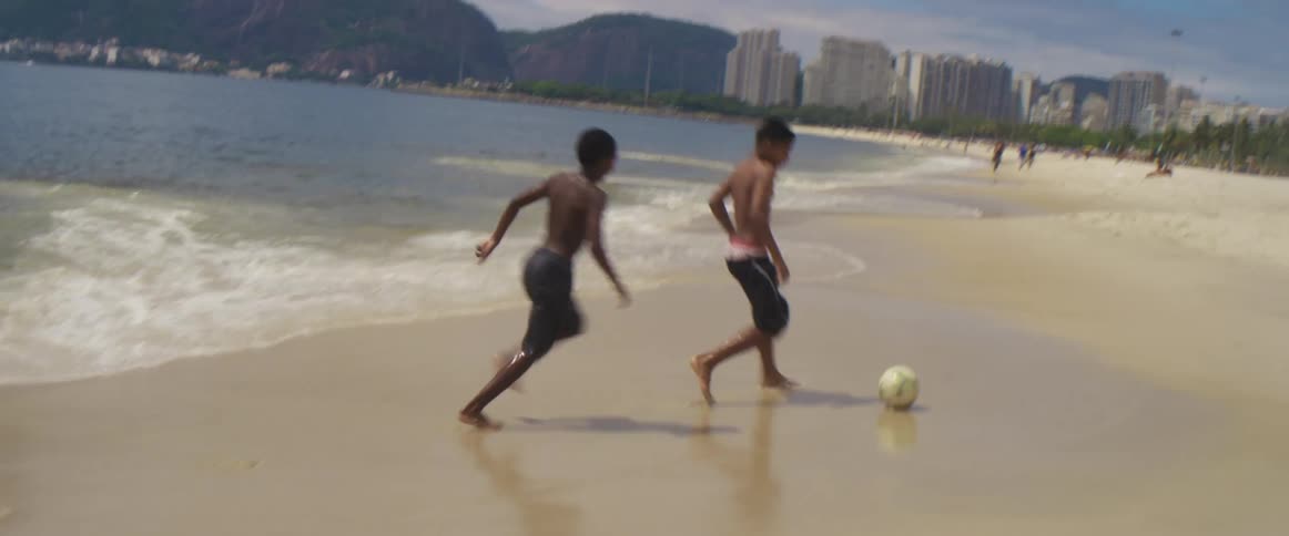 A group of youths plays football on the white sand beach next to the ocean
