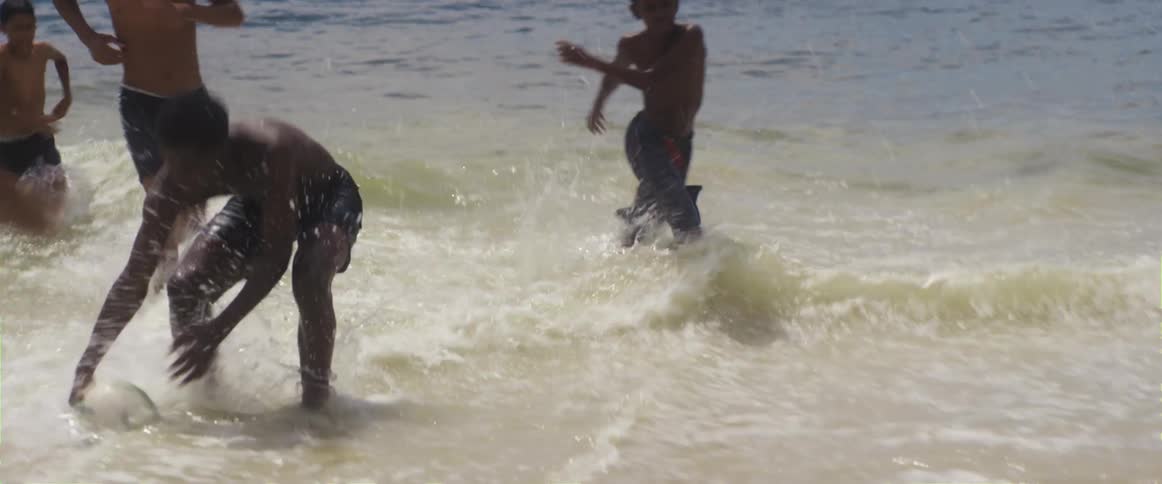 Two boys kick a ball on a beach and a group of boys run in from the water to pick up the ball and kick it back to them
