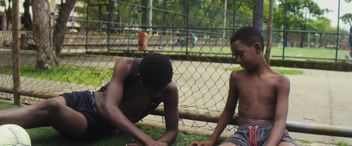 A teenage boys sits on the grass with a soccer ball and talks to a young boy as people walk behind them