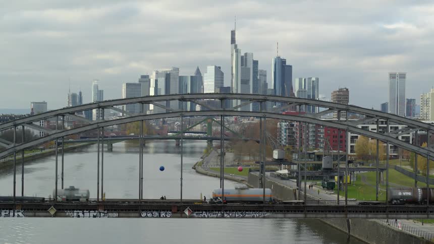 Train and traffic cross a series of bridges across river with city skyline in distance