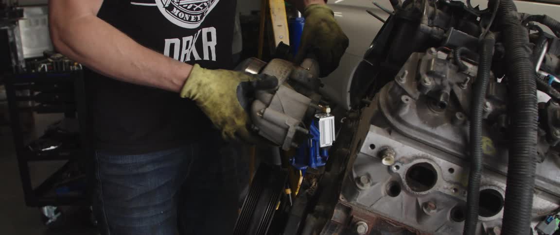 Mechanic works on car engine sitting outside of car as another man walks past nearby car