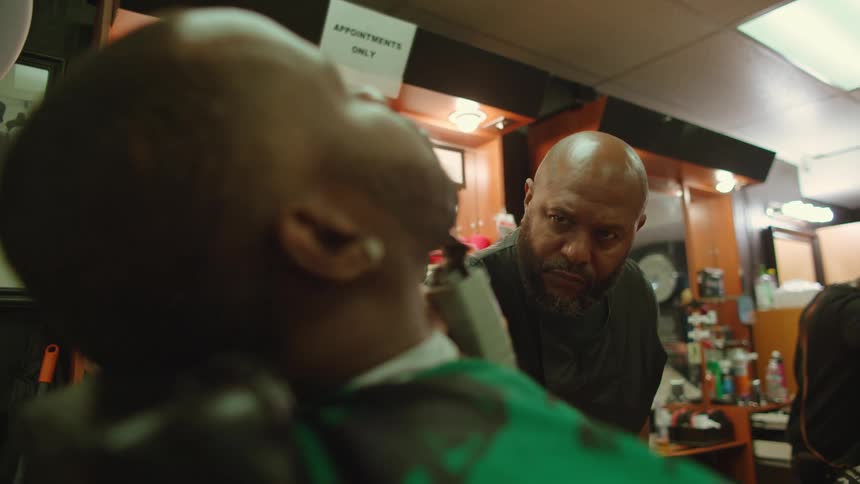 A man sits in a chair with his head back as a barber shaves his beard