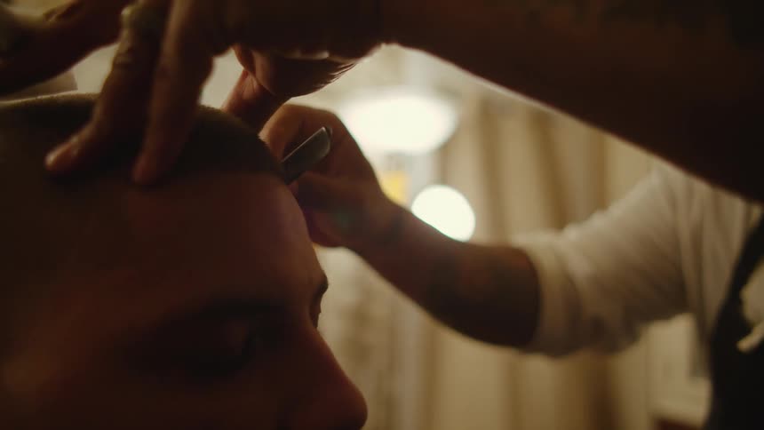 Barber with white hoodie and black apron cuts mans hair with straight razor in barber shop