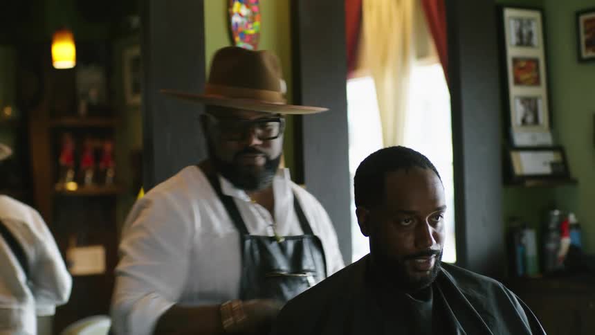 Barber wearing a hat combs the beard of a talking client sitting on a barber shop