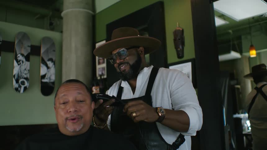Barber with brown hat glasses beard white shirt and black apron cuts mans hair with clippers in barbers shop