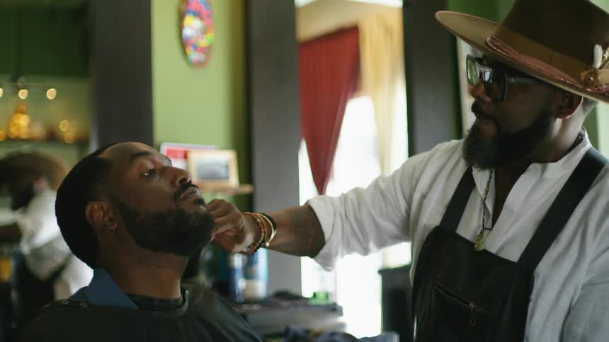 A man sitting in a barbers chair lifts his head as the barber combs his beard